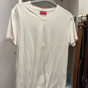 Vit t-shirt från Hugo Boss - Säljer en stilren vit t-shirt från Hugo Boss med v-ringning. Perfekt basplagg för alla tillfällen. T-shirten är kortärmad och gjord i mjukt material för extra komfort.