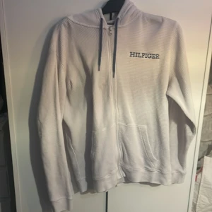 Vit hoodie från Tommy Hilfiger - Säljer en stilren vit hoodie från Tommy Hilfiger med dragkedja och broderad logga på bröstet. Tröjan har långa ärmar och en bekväm huva med snörning. Pris kan diskuteras 