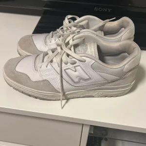 Vita sneakers från New Balance - Säljer ett par stilrena vita New Balance 550 sneakers med grå detaljer. Skorna har snörning och är tillverkade i en kombination av skinn och syntetmaterial. Perfekta för en avslappnad stil.