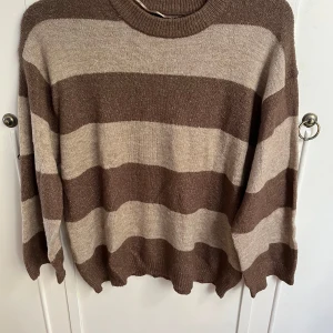 Brun och beige randig tröja från 157 - Säljer en mysig brun och beige randig tröja från 157. Tröjan har långa ärmar och en rund halsringning, perfekt för kyligare dagar. Den är tillverkad i ett mjukt material som känns skönt mot huden.