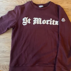 Vinröd sweatshirt med St Moritz-tryck - Snygg vinröd sweatshirt från Moncler med St Moritz-tryck i vitt på bröstet. Tröjan har långa ärmar och en rund halsringning. Perfekt för en avslappnad stil med en touch av lyx. Storlek M, möts i Solna