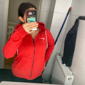 Röd hoodie med dragkedja från Nike - Säljer en röd hoodie från Nike med dragkedja och vit logga på bröstet. Den har en justerbar huva med vita snören och två fickor framtill. Perfekt för en sportig look eller en avslappnad dag.
