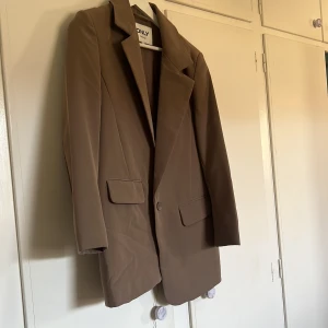 Brun kavaj från ONLY - Snygg brun kavaj från ONLY med klassisk design. Kavajen har en knapp framtill och två fickor. Endast använd ett fåtal gånger så mycket fint skick! 