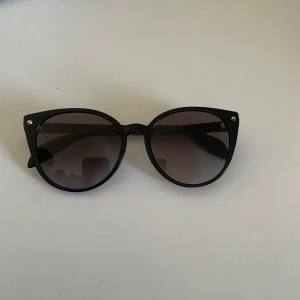 Svarta solglasögon från Alexander McQueen - Snygga svarta solglasögon från Alexander McQueen med rundad form och subtila gulddetaljer vid gångjärnen. Perfekta för en stilren look.