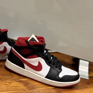 Nike Air Jordans i svart, vitt och rött (Gym Red) - Snygga Nike Air Jordans i en klassisk färgkombination av svart, vitt och rött. Skorna har en ikonisk design med snörning och den välkända swoosh-loggan i rött på sidan. Perfekta för dig som vill ha en stilren och sportig look.