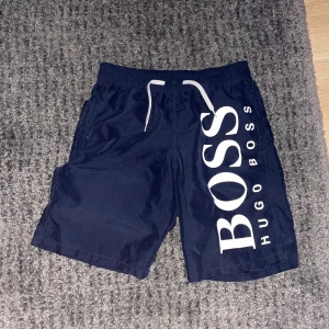 Marinblå badshorts från Hugo Boss - Snygga marinblå badshorts från Hugo Boss med stor vit logga på sidan. De har en elastisk midja med snörning för justerbar passform. Perfekta för sommarens stranddagar.