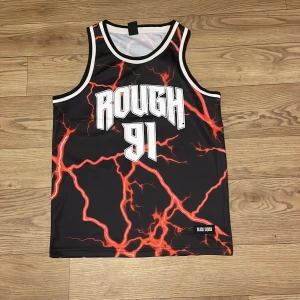 Svart linne från Black Squad med blixtmönster - Coolt svart linne från Black Squad med ett iögonfallande rött blixtmönster och texten 'ROUGH 91' på framsidan. Perfekt för en sportig look eller som statement-plagg. Tillverkad i ett lätt och bekvämt material.