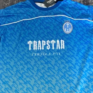 Blå fotbollströja  - Snygg blå fotbollströja  med ett unikt mönster och texten 'IT'S A SECRET' på framsidan. Tröjan har korta ärmar och nummer 22 på baksidan. Perfekt för dig som vill sticka ut på planen eller gatan.