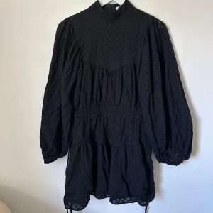 Svart broderad kortklänning Shona Joy - Supersnygg svart kortklänning från Shona Joy med broderade detaljer och hög hals. Klänningen har långärmade puffärmar, markerad midja och volangkant nedtill. Tillverkad i 100% bomull för en skön och luftig känsla. Perfekt för dig som gillar en chic och trendig look. B356