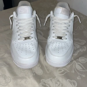 Nike Air Force 1 helvita sneakers - Klassiska Nike Air Force 1 sneakers i helvitt skinn med vit sula och snörning. Ikonisk design med perforerad tå och Nike-logga bak. Perfekta för dig som vill ha en clean och tidlös look till vardags. Storlek 44 (US 10, UK 9). Pris kan diskuteras