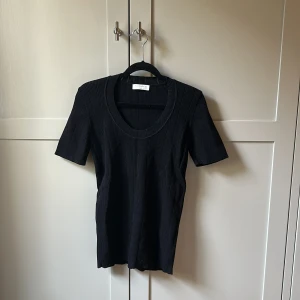 Svart ribbad topp från Sandro Paris - En svart ribbad topp från Sandro Paris med korta ärmar och rund halsringning. Toppen har en slim passform och är tillverkad i ett stretchigt material som sitter snyggt på kroppen. Perfekt att styla med jeans eller kjol för en clean look.