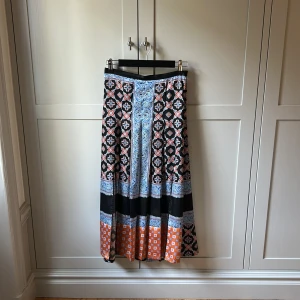 Sandro långkjol i svart och blått - Säljer en maxilång kjol med coolt grafiskt mönster i svart, blått, orange och vitt. Kjolen har breda bårder med detaljerade tryck och en flowy passform nertil, och kroppsnära upptill. Perfekt för dig som gillar färg och unika prints. Materialet känns lätt och mjukt mot huden. Sandro storlek ”1”