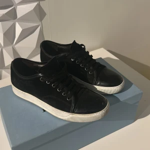 Svarta sneakers från Lanvin i mocka - Snygga svarta sneakers från Lanvin med ovandel i mocka och detaljer i skinn. Skorna har rund och glansig tå, svarta snören och vit platt sula. Perfekta för dig som gillar stilrena och lyxiga sneakers med en clean look.