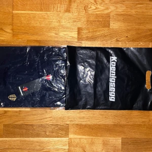 Mörkblå pikétröja från Koenigsegg - Snygg mörkblå pikétröja från Koenigsegg med klassisk krage och knappar framtill. Tröjan har ett broderat Koenigsegg-emblem på bröstet och är tillverkad i mjuk bomull. Perfekt för dig som gillar exklusiva detaljer och stilren design. Tröjan är köpt i Ängelholm alla lappar och original tillbehör sitter kvar och följer självklart med i affären. XS men passar som S.