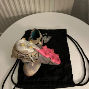 Puma beige fotbollsskor utan snörning. Elite - Säljer ett par grymma Puma fotbollsskor i beige med inslag av vitt och svart, samt coola rosa detaljer på sulan. Skorna är utan snörning och har en strumpliknande passform för bästa känsla på planen. + en skobag från Nike på köpet.