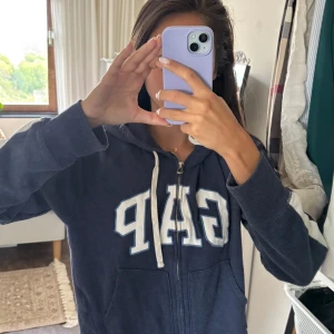 Blå GAP hoodie med dragkedja - Mörkblå hoodie från GAP med vit logga framtill och vita detaljer på ärmarna. Tröjan har huva med snörning, dragkedja och två fickor framtill. Perfekt för chill dagar och enkel att matcha med jeans eller leggings.