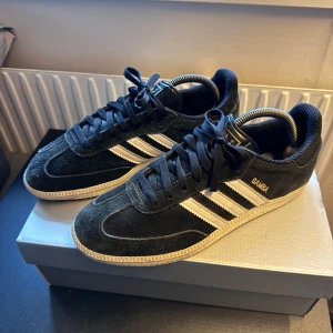 Adidas Samba svarta sneakers i mocka - Klassiska Adidas Samba sneakers i svart mocka med vita ränder och gummisula. Skorna har snörning och rund tå, samt den ikoniska SAMBA-loggan i guld på sidan. Perfekta för dig som gillar retrostil och streetwear. Lite korta snören