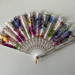 Blommig solfjäder med spetskant - Snygg solfjäder med färgglatt blommönster i lila, rosa, blått och grönt. Spetskant upptill och vit plast med gulddetaljer på handtaget. 