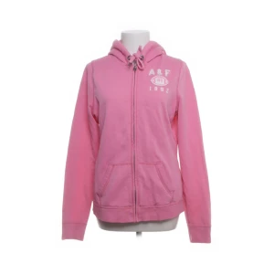 Rosa hoodie från Abercrombie & Fitch zip up  - Snygg rosa hoodie från Abercrombie & Fitch med dragkedja, huva med snörning och vita tryckdetaljer på bröstet. Perfekt för chill dagar eller när du vill ha en avslappnad look. Mjuk bomullskänsla och kängurufickor framtill.