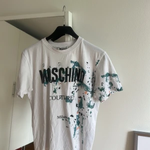 Vit Moschino Couture t-shirt med tryck - Snygg vit t-shirt från Moschino Couture med coolt färgstänksmönster i grönt och orange över hela framsidan. Stor logotyp och text på bröstet. Klassisk rund hals och korta ärmar. Perfekt statement-plagg för dig som gillar designer och streetstyle.