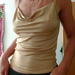 Champagnefärgad satin linnetopp - glansig champagnefärgad linnetopp med smala axelband och snygg drapering vid bysten. Figursydd o fin passform