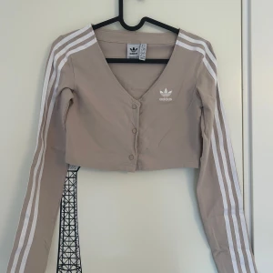 Beige croppad topp från Adidas - Beige croppad topp med vita ränder längs ärmarna och v-ringning från Adidas i storlek XS. Toppen har lång ärm, tryckknappar framtill och Adidas-logga på bröstet. Aldrig använd. 