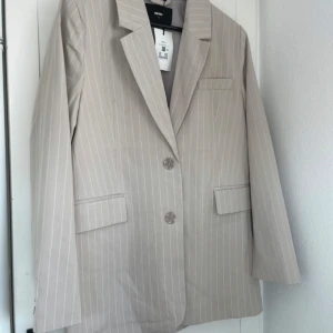 Beige randig kavaj med knappar - Snygg beige kavaj med tunna vita ränder och två knappar framtill. Klassisk krage och fickdetaljer, perfekt för dig som gillar en stilren look. Passformen är normal och materialet känns lätt och bekvämt. Enkel att matcha med jeans eller kostymbyxor.