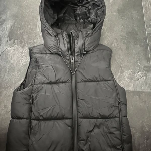 Svart dunväst med huva från Zara - Svart dunväst från Zara med stor, vadderad huva och snörning. Västen har quiltad design, dragkedja framtill och justerbar dragsko vid halsen. Perfekt för kyliga dagar när du vill ha en clean och sportig look.