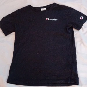 Svart Champion t-shirt i bomull - Svart t-shirt från Champion med klassisk logga broderad på bröstet och ärmen. Skön passform och rund halsringning, perfekt till jeans eller shorts. Tillverkad i mjuk bomull som andas. Enkel och stilren design som funkar året runt.