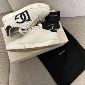 Dolce & Gabbana vita sneakers med svart logo - Snygga vita sneakers från Dolce & Gabbana med stor svart logga på sidan och svart detalj vid hälen. Skorna har snörning och är tillverkade i skinn. Perfekta för dig som vill ha en clean och lyxig look med streetkänsla.