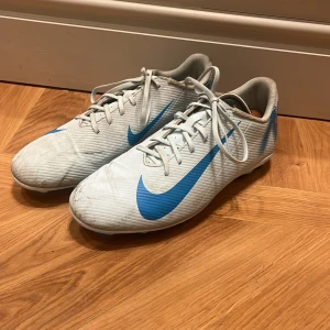 Nike Mercurial fotbollsskor - Nike Mercurial fotbollsskor i vitt med blå detaljer och stor Nike-logga på sidan och hälen. Skorna har snörning, låg profil och är tillverkade i syntetmaterial för bästa grepp på planen. Perfekta för dig som vill sticka ut med stil på matchen.