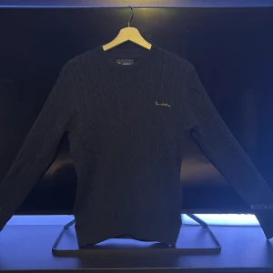 Mörkblå kabelstickad tröja Bondelid - Säljer en mörkblå kabelstickad tröja från Bondelid med broderad logga på bröstet. Tröjan har rund halsringning och långa ärmar, perfekt för lager-på-lager. Klassisk och stilren design som funkar till många outfits. Snygg till vintern då den är stickad o lite mönstrad, hör av dig!!