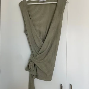 Ribbad omlottopp från Mango - Snygg ribbad omlottopp från Mango i en ljusbeige nyans. Toppen är ärmlös, har djup v-ringning och knyt i sidan för en cool och avslappnad look. Perfekt att styla över en skjorta eller t-shirt för en trendig layering-effekt.