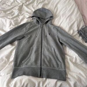 Grå hoodie från Polo Ralph Lauren - Säljer en grå hoodie från Polo Ralph Lauren med dragkedja och huva. Tröjan har två fickor fram, vita snören och den klassiska lilla Polo-loggan broderad på bröstet. Perfekt för chill dagar och enkel att matcha med jeans eller joggers. Strl xs passar även M. Pris kan diskuteras