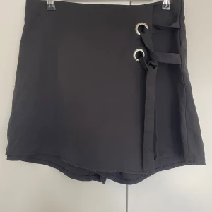 Svart omlotts skorts med snörning - Svarta skorts med omlott-design och snygga snörningsdetaljer med metallringar på sidan. Skortsen har en stilren look och är gjord i ett mjukt tyg som känns skönt mot huden. Perfekt för dig som gillar minimalistisk stil med en twist.