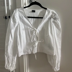 Vit blus med knyt från Gina tricot - Säljer en vit blus från Urban Bliss med v-ringning och knyt framtill. Blusen har långa puffärmar med resår vid ärmslut och en luftig, lite croppad passform. Perfekt för dig som gillar en romantisk och trendig stil.