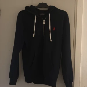 Svart hoodie från Polo Ralph Lauren - MARIN BLÅ  hoodie från Polo Ralph Lauren med vit dragsko och dragkedja framtill. Klassisk röd broderad logga på bröstet. Mjuk bomullskänsla och huva. Perfekt för en chill och stilren look.