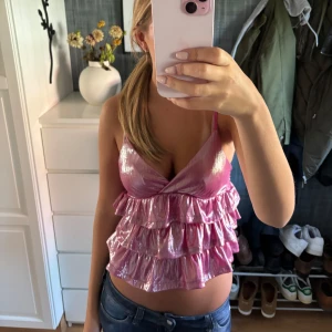 Glittrig rosa volangtopp - Säljer en glittrig rosa topp med smala axelband och flera lager volanger. Toppen har omlott-skärning framtill och är croppad, perfekt för dig som vill sticka ut. Materialet är glansigt och fångar ljuset snyggt.