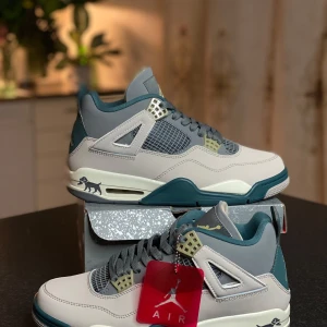 Nike Air Jordan 4 Retro grå/blå - Nike Air Jordan 4 Retro sneakers i grått och blågrönt med vita och beige detaljer. Skorna har meshpaneler, snörning och Jumpman-logga på häl och sula. Ovandelen är i skinn och syntet, med cool hunddetalj på sidan av sulan. Perfekta för dig som gillar streetstyle.