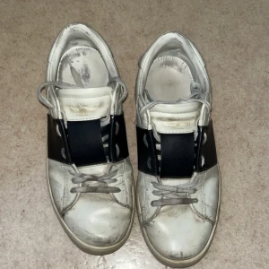 Svarta Valentino opens  - Säljer ett par vita sneakers från Valentino Garavani med svart bred rem över mitten. Skorna har snörning, rund tå och platt sula. Tillverkade i skinn och har en stilren design med diskreta detaljer. Perfekta för dig som gillar lyxiga och trendiga sneakers. Storlek 43 helt okej skick bra pris vid snabb affär 