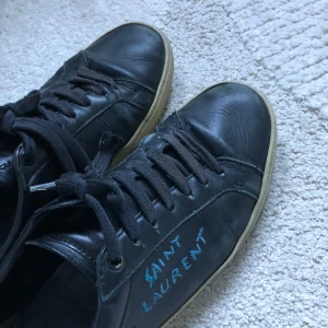 Svarta Saint Laurent sneakers i skinn - Svarta sneakers från Saint Laurent med handskriven logga i blått på sidan. Skorna har rund tå, svarta snören och en vit platt sula. 