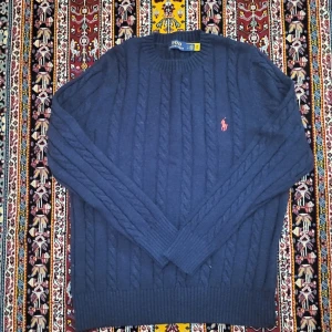 Mörkblå kabelstickad tröja Polo Ralph Lauren - Kabelstickad mörkblå tröja från Polo Ralph Lauren med klassisk rund hals och broderad rosa logga på bröstet. Tröjan har långa ärmar och ribbade muddar för en snygg passform. Perfekt för dig som gillar stilren och tidlös design.