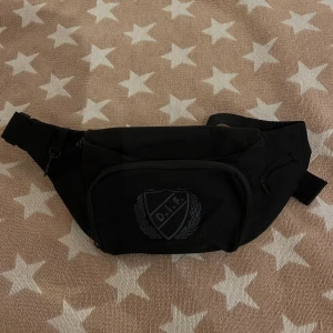 Svart magväska med DIF-logga - Säljer en svart magväska med broderad DIF-logga framtill. Väskan har flera fack med dragkedja och justerbart band för midjan. Perfekt för att bära det viktigaste när du är på språng.