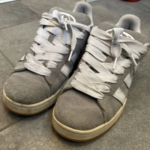 Grå adidas Campus sneakers i mocka - Snygga adidas Campus sneakers i grå mocka med vita detaljer och klassiska tre ränder på sidan. Skorna har vit snörning, rund tå och en gummisula i ljusbrun färg. Perfekta för dig som gillar retrovibbar och streetstyle.