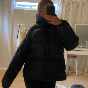 Svart pufferjacka med huva - Säljer en svart pufferjacka med huva. Jackan har en dragkedja framtill och är perfekt för kyliga dagar. Den är designad för att hålla dig varm och bekväm med sin vadderade stil.