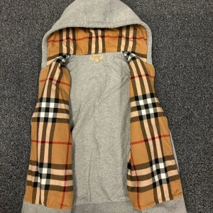 Grå hoodiejacka från Burberry - Grå hoodiejacka från Burberry med klassiskt rutigt foder i beige, svart, vitt och rött. Jackan har huva och är tillverkad i mjuk bomull. Perfekt för dig som vill ha en stilren look med en touch av lyx.