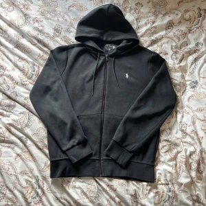 Svart hoodie från Polo Ralph Lauren - Svart hoodie från Polo Ralph Lauren med dragkedja och huva. Klassisk design med vit broderad logga på bröstet, snörning i huvan och två fickor framtill. Tillverkad i mjuk bomull för en skön och avslappnad stil.