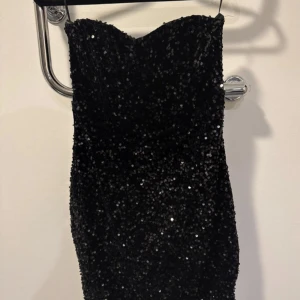 Svart glittrig kort klänning från Amisu - Supersnygg svart axelbandslös klänning från Amisu, täckt med glittriga paljetter som ger en riktigt festlig vibe. Modellen är kort och har en tight passform som framhäver figuren. Perfekt för dig som vill sticka ut med lite extra bling.