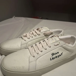 Vita Saint Laurent sneakers - Snygga vita sneakers från Saint Laurent med handskriven logga på sidan. Skorna har klassisk låg profil, rund tå och platt sula. Tillverkade i canvas och skinn med vita snören och diskreta detaljer. Perfekta för dig som gillar stilrena och exklusiva sneakers.