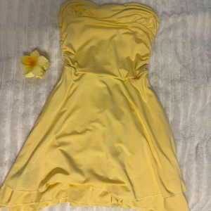 Gul axelbandslös kortklänning - Säljer en söt gul axelbandslös klänning med lätt rynk vid bysten och mjukt fall. Klänningen har en kort längd och är perfekt för soliga dagar. Materialet känns lätt och bekvämt mot huden. 🌼